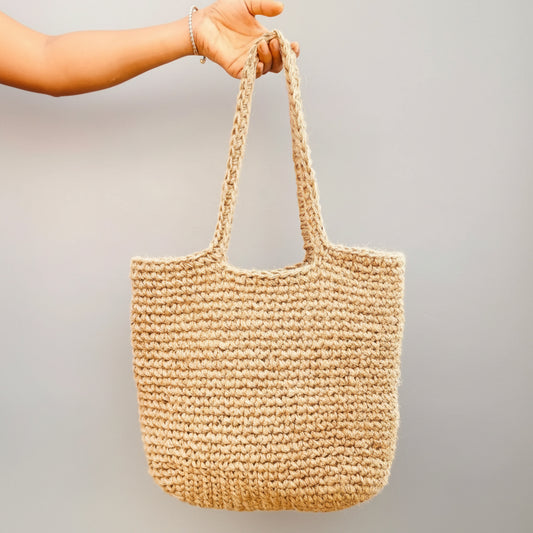 Hand holding a woven beige bag with green plants in the background