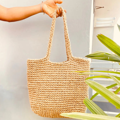 Hand holding a woven beige bag with green plants in the background
