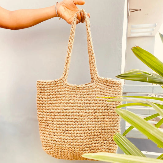 Hand holding a woven beige bag with green plants in the background