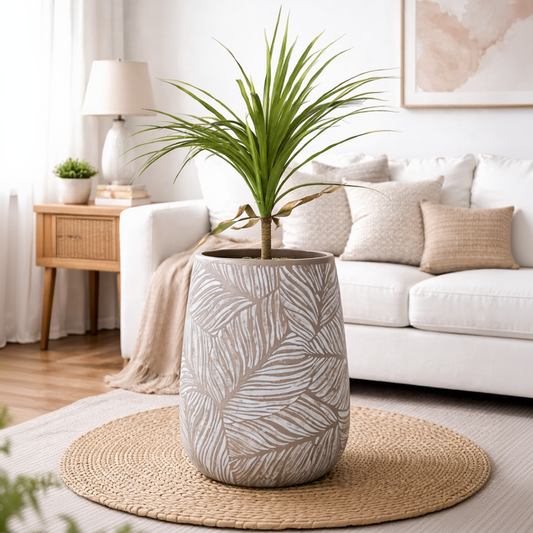 A tall cylindrical planter with a gray and white leaf pattern, holding a green palm plant, placed on a round woven rug in a bright, modern living room.